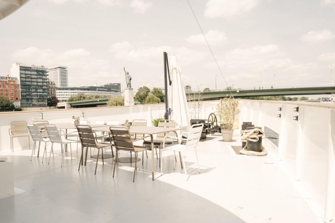 Terrasse extérieure d'un bateau avec des tables et chaises, vue sur un pont, des bâtiments et une statue au loin.