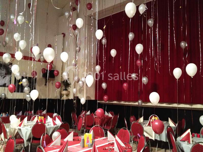 Salle décorée pour un événement avec des ballons rouges, blancs et argentés suspendus au plafond et des tables dressées.