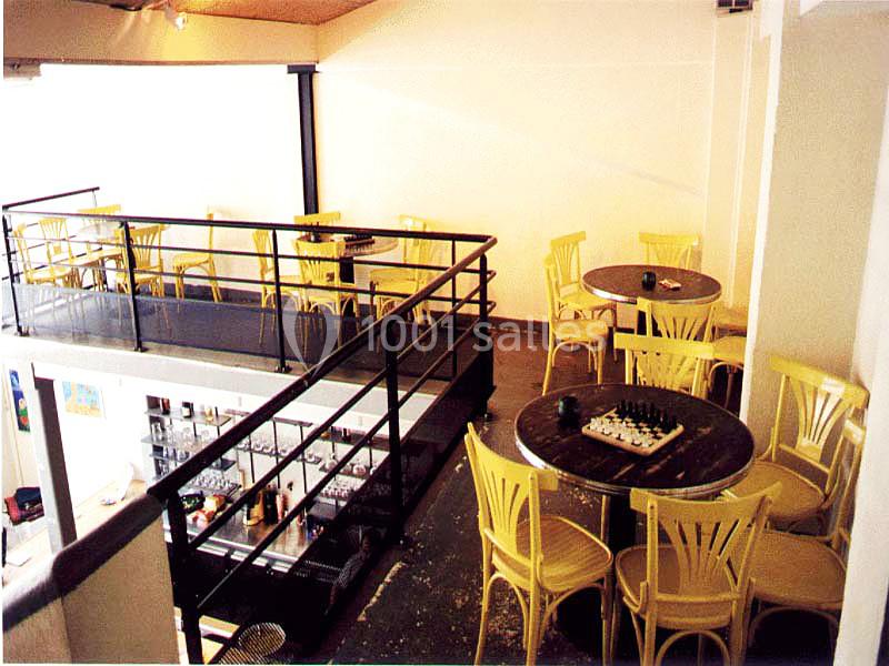 Salle de café avec mezzanine, tables rondes en bois, chaises jaunes et un échiquier posé sur une table.