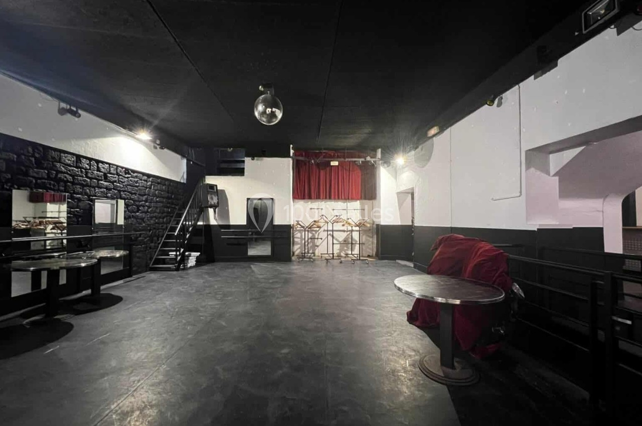 Salle vide avec sol noir, murs blancs et noirs, rideaux rouges au fond, tables et chaises empilées.