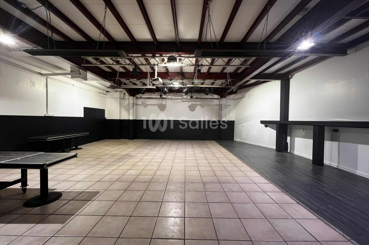 Salle vide avec sol en carrelage et parquet, murs blancs et noirs, plafond avec poutres apparentes et éclairage suspendu.