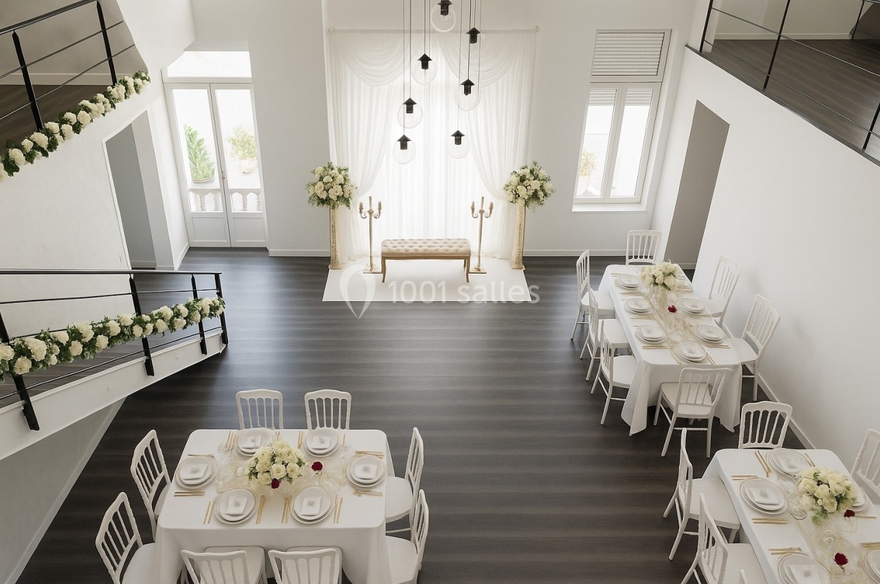 Salle lumineuse décorée pour un mariage, avec tables dressées, fleurs blanches et espace pour une cérémonie.