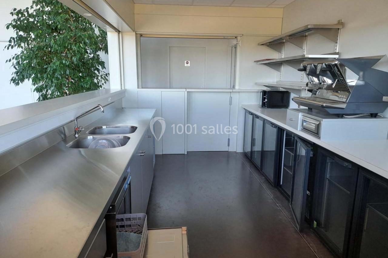 Cuisine professionnelle avec plan de travail en inox, évier double, étagères murales, machines à café et réfrigérateurs.