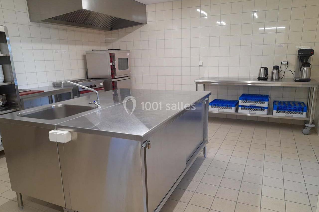 Cuisine professionnelle avec plan de travail en inox, évier, étagères, four, cafetière et carrelage mural blanc.