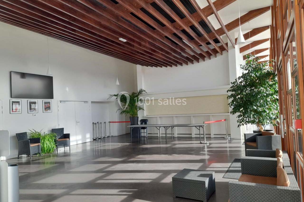 Hall lumineux avec grandes baies vitrées, plafond en bois, mobilier d'attente et plantes décoratives.