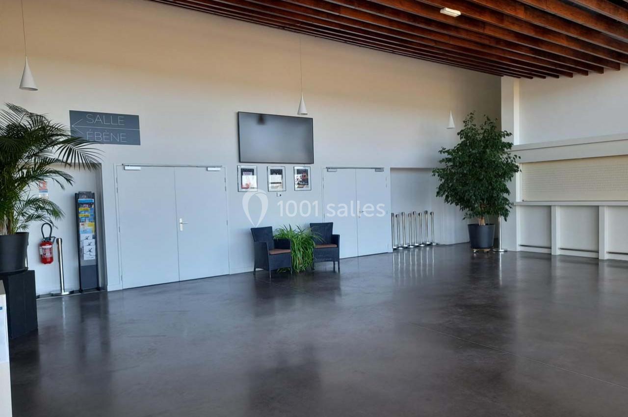 Hall lumineux avec sol en béton, plafond en bois, plantes en pot, chaises, et portes blanches sous un écran mural.