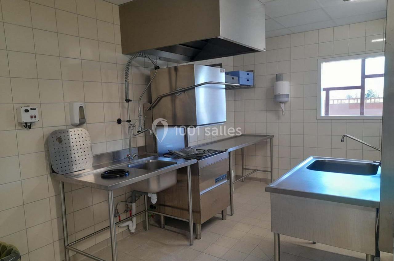 Cuisine professionnelle avec évier, lave-vaisselle industriel, plans de travail en inox et carrelage mural clair.