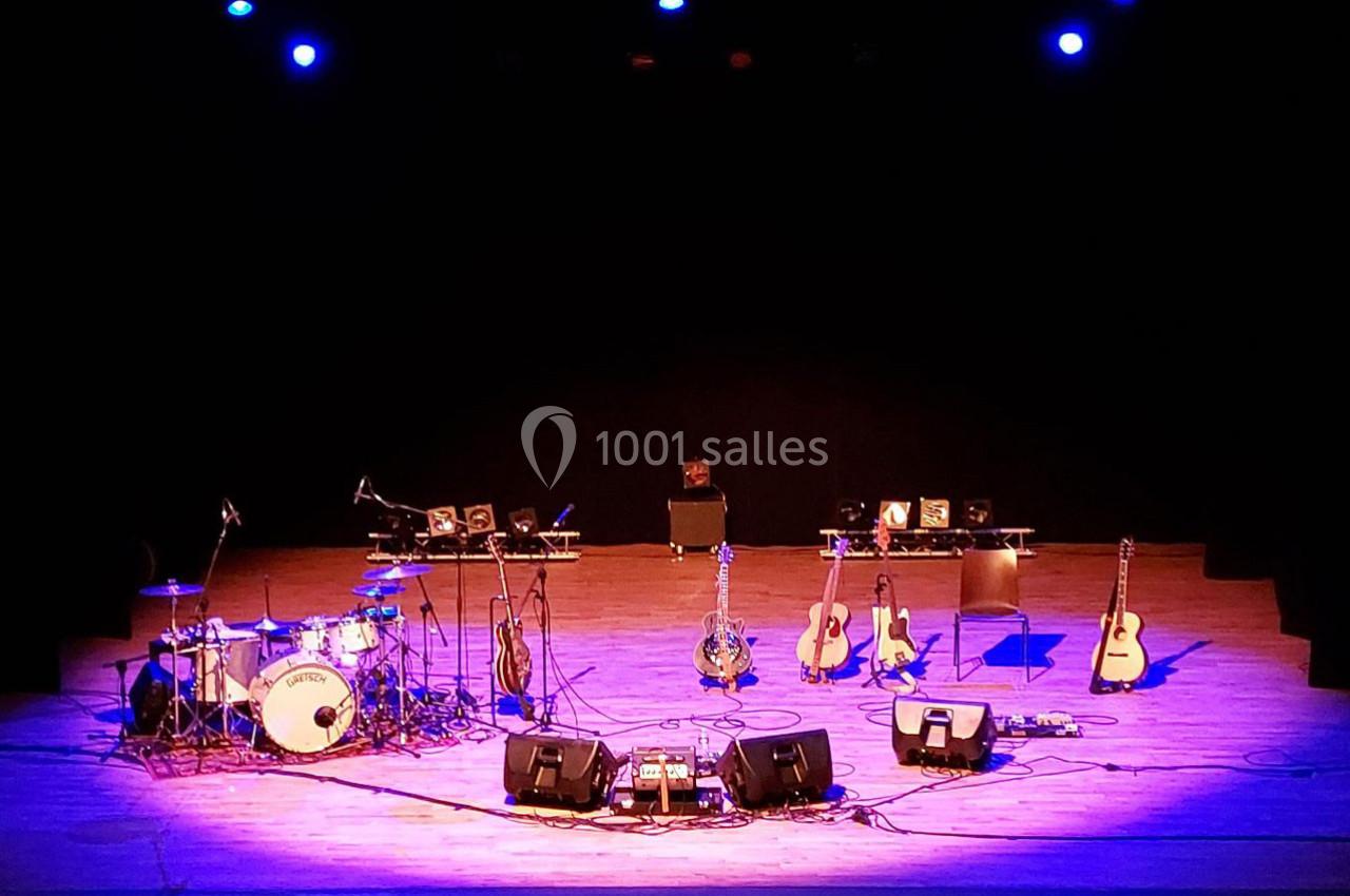 Scène de concert vide avec des instruments de musique, dont des guitares, une batterie et des micros, sous un éclairage bleu.
