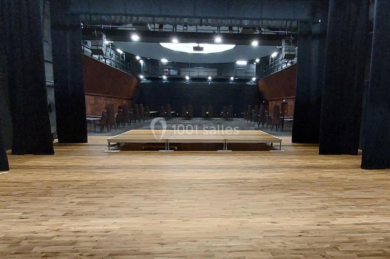 Salle de spectacle vide avec une scène en bois au centre, entourée de rideaux noirs et de rangées de sièges.