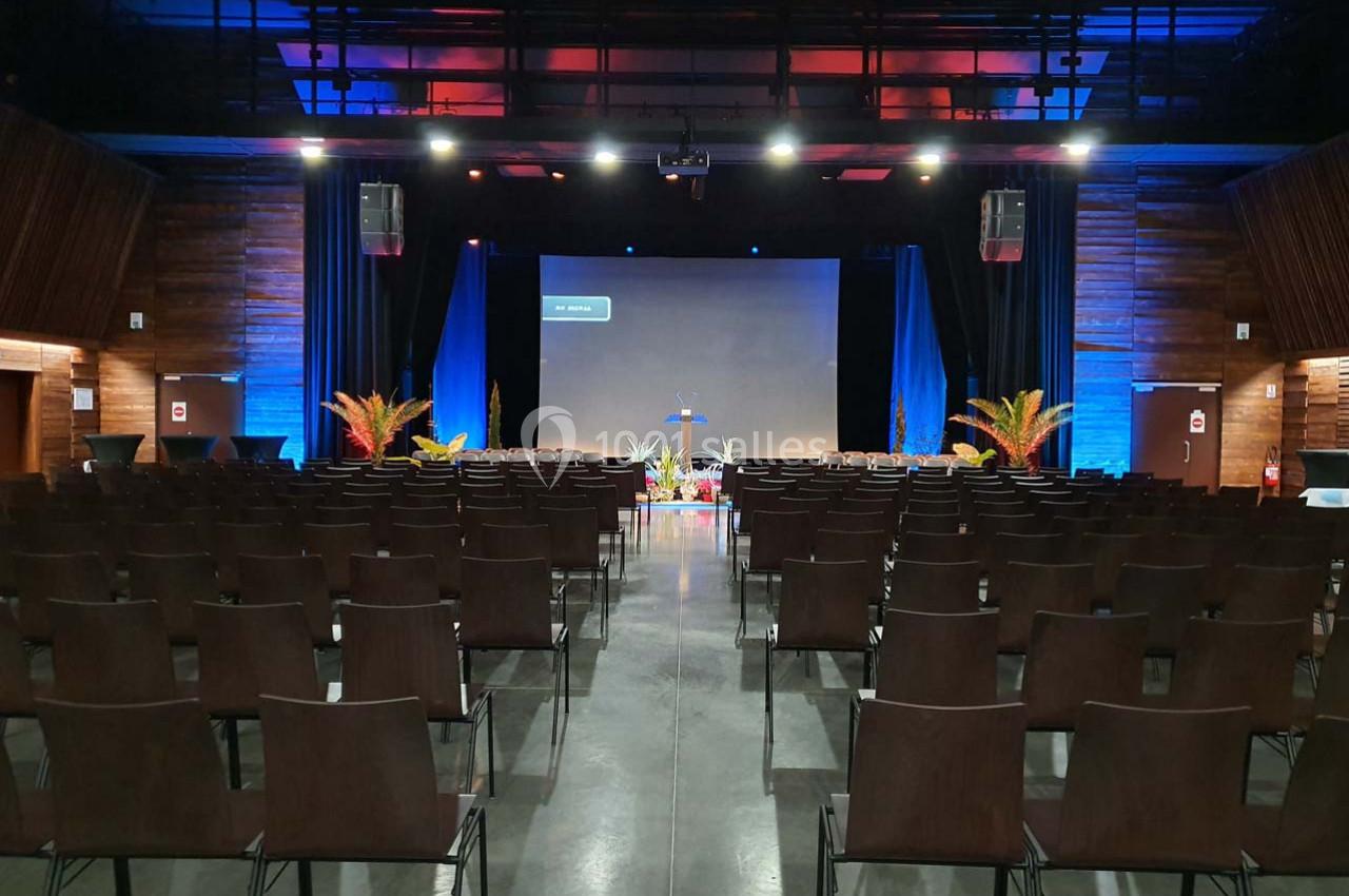 Salle de conférence vide avec des rangées de chaises, un écran de projection et une scène décorée de plantes.