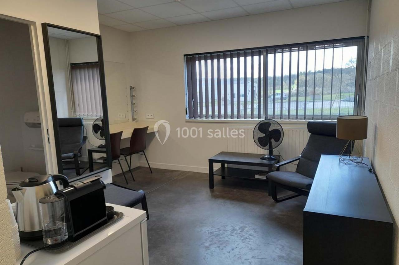Salle lumineuse avec un bureau, fauteuils, miroir, cafetière et fenêtre à stores donnant sur un extérieur verdoyant.