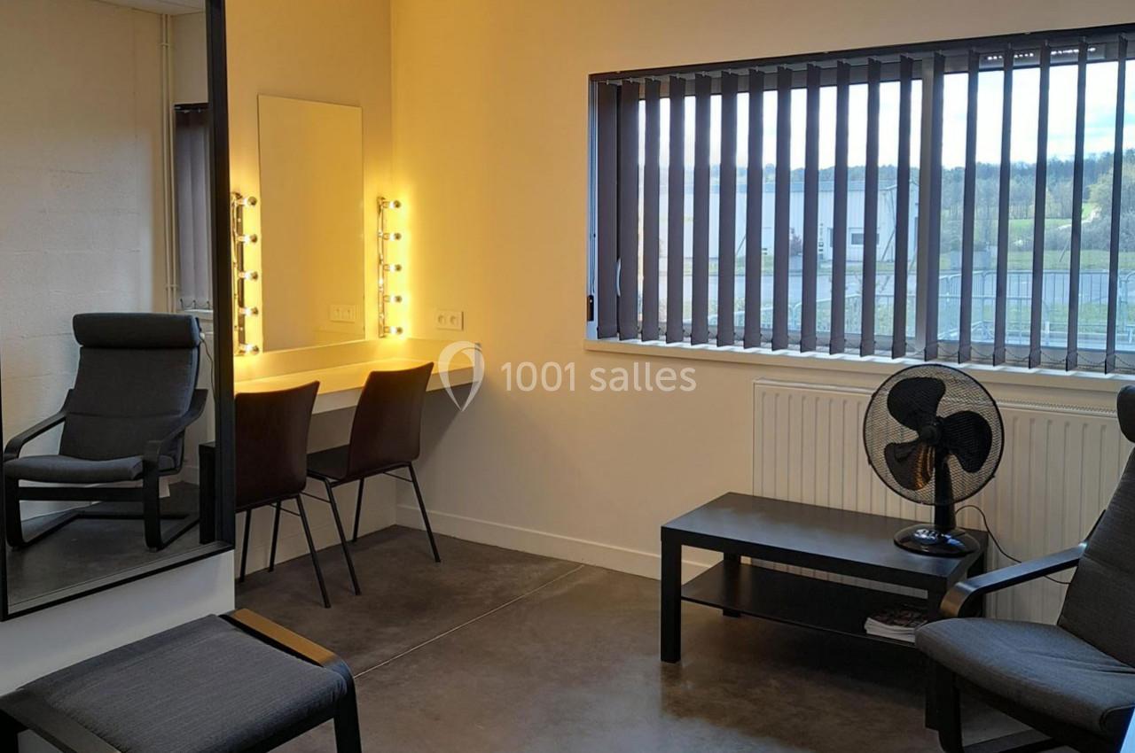 Salle de loge avec miroir éclairé, chaises, table basse, ventilateur et fenêtre avec stores.