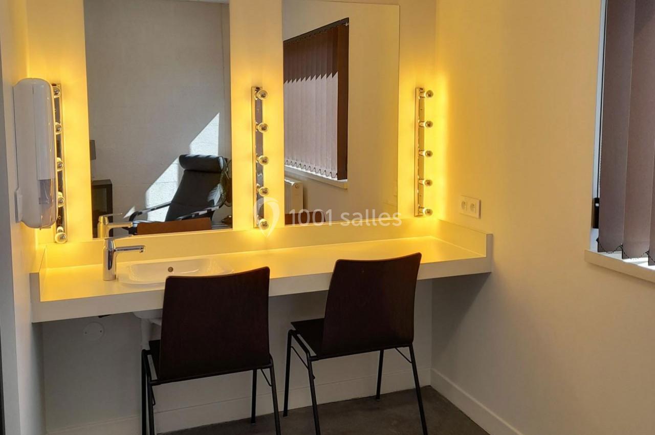 Coiffeuse avec miroir éclairé par des ampoules, deux chaises noires et un lavabo intégré dans une pièce lumineuse.