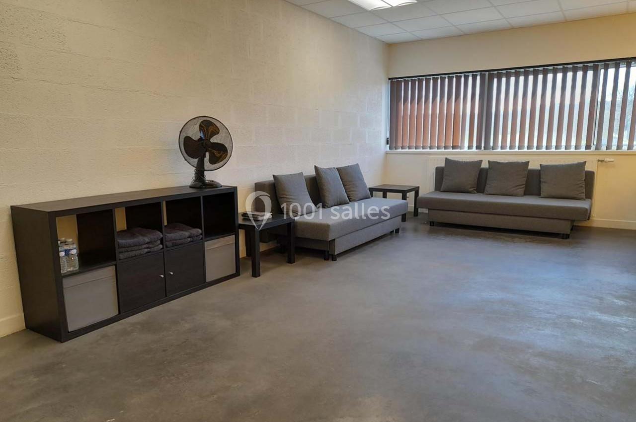 Salle lumineuse avec canapé gris, étagère noire, ventilateur vintage et sol en béton poli.