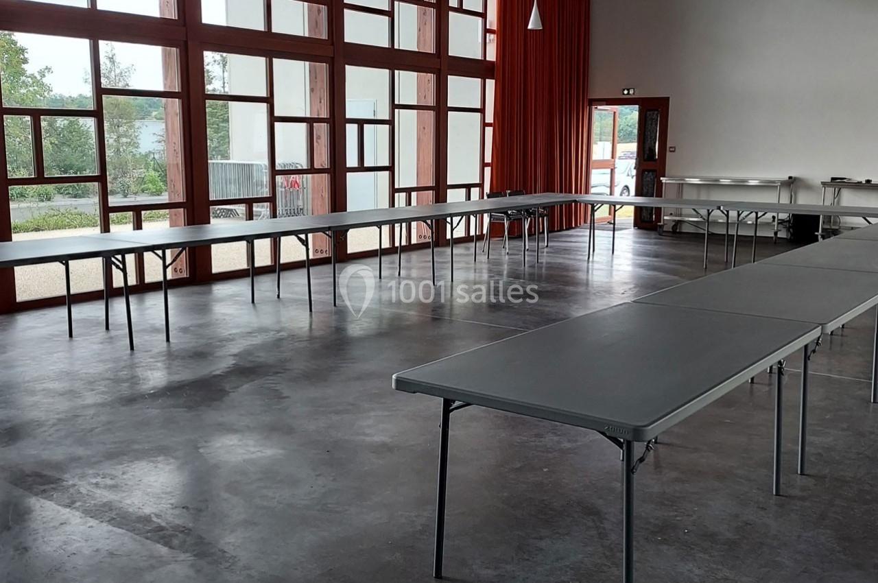 Salle lumineuse avec de grandes baies vitrées, tables pliantes grises disposées en U sur un sol en béton.