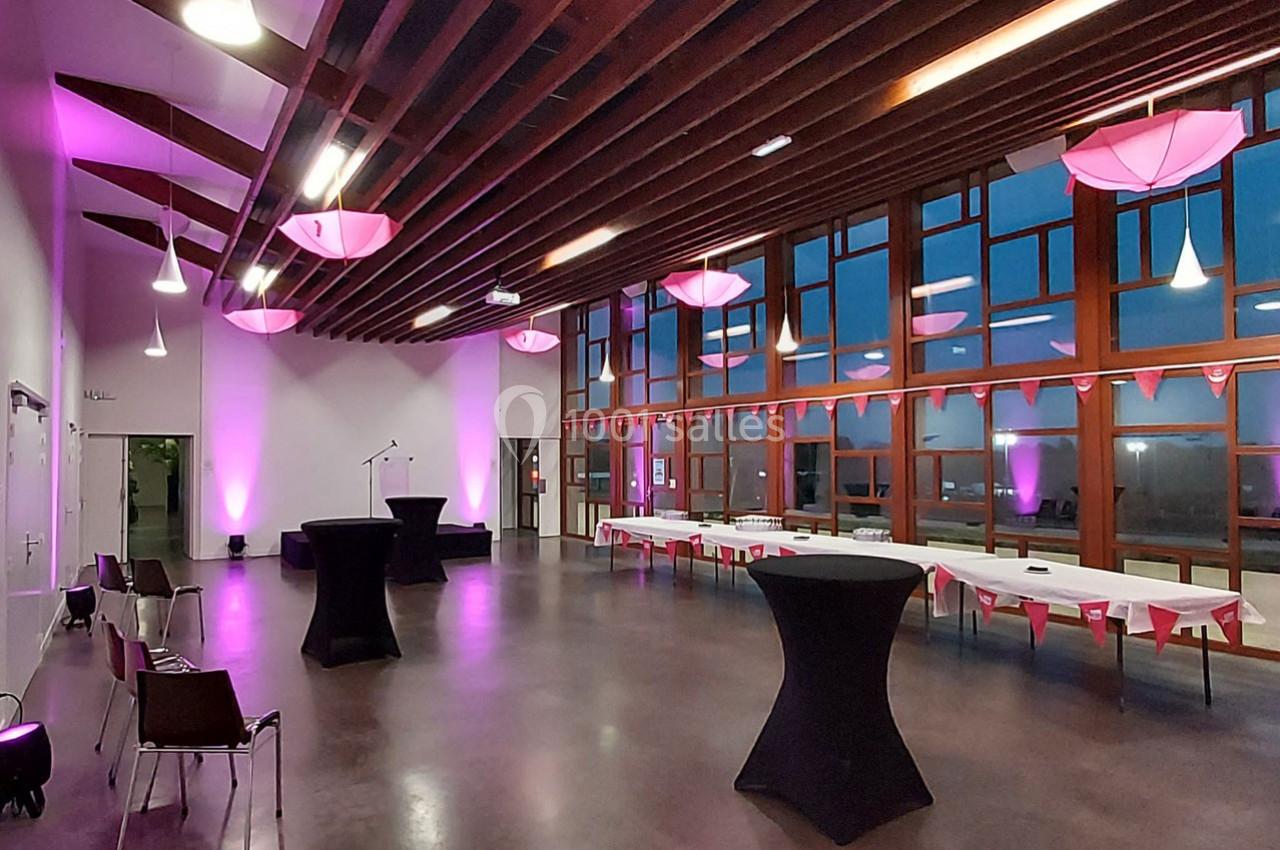Salle de réception avec tables décorées, chaises, éclairage rose et grandes baies vitrées donnant sur l'extérieur.