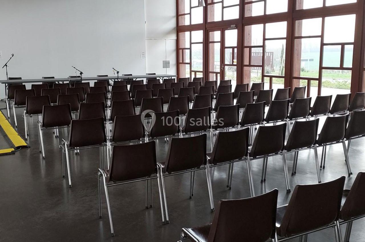 Salle de conférence vide avec des rangées de chaises marron, des micros sur une table et de grandes fenêtres vitrées.