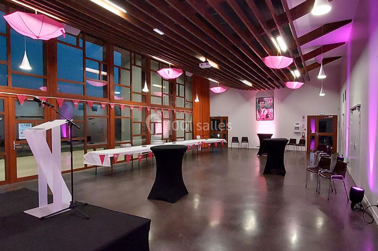 Salle lumineuse avec grandes baies vitrées, tables dressées, chaises alignées et éclairage rose pour un événement.