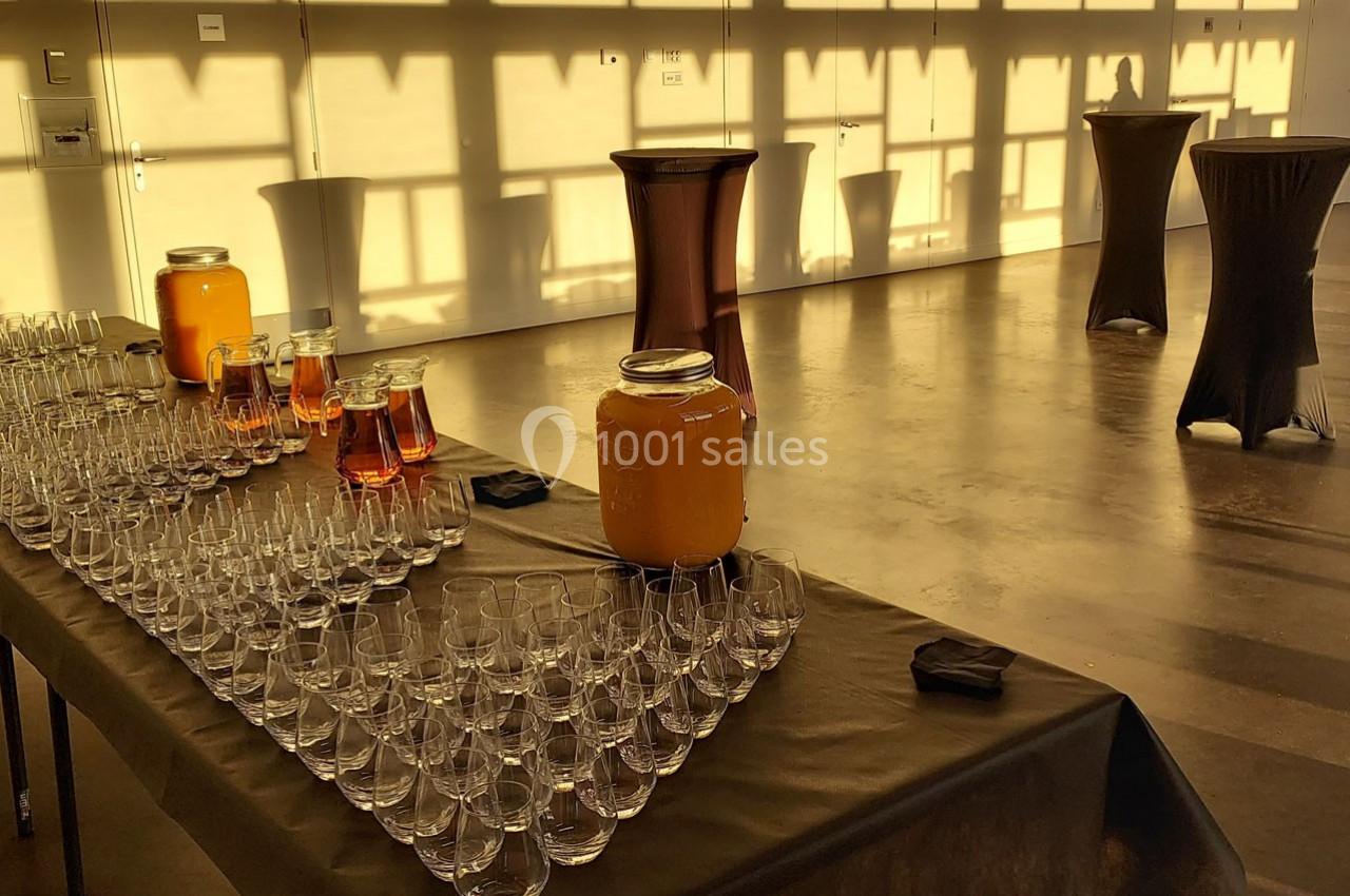 Salle lumineuse avec des tables hautes et une table dressée contenant des verres, des carafes et des boissons.