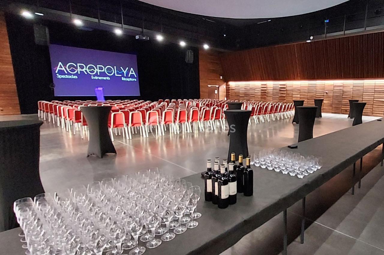 Salle de conférence avec rangées de chaises rouges, scène éclairée et table préparée avec verres et bouteilles de vin.