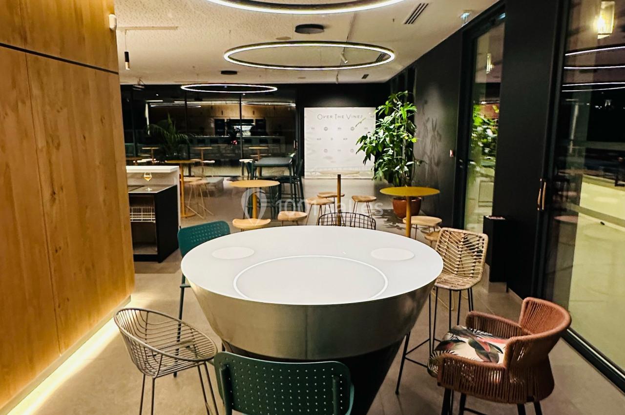Salle moderne avec table ronde centrale, chaises variées, éclairage circulaire suspendu et grandes baies vitrées.