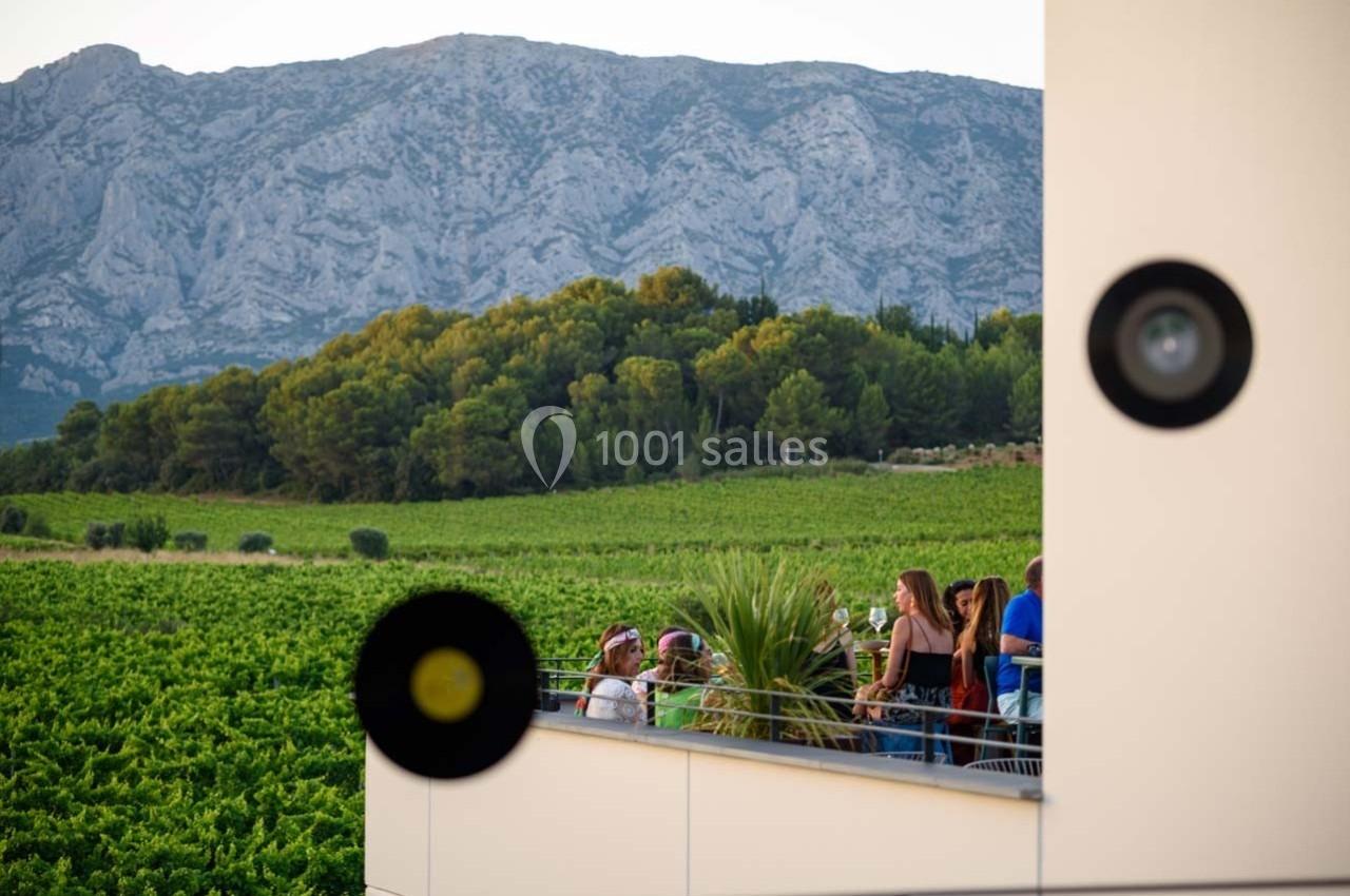 Des personnes discutent sur une terrasse surplombant un vignoble verdoyant avec des montagnes en arrière-plan.