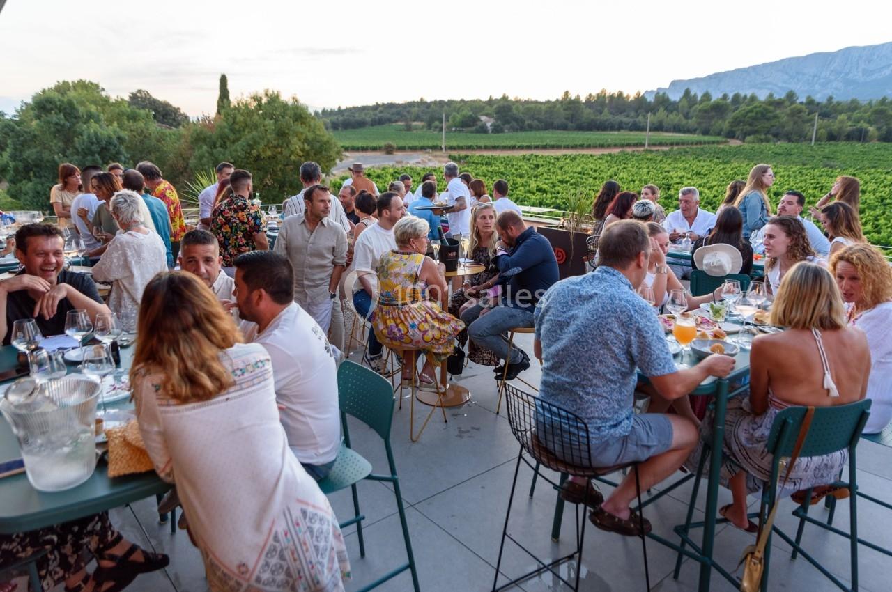 Groupe de personnes dînant en terrasse avec vue sur des vignobles et des collines au coucher du soleil.