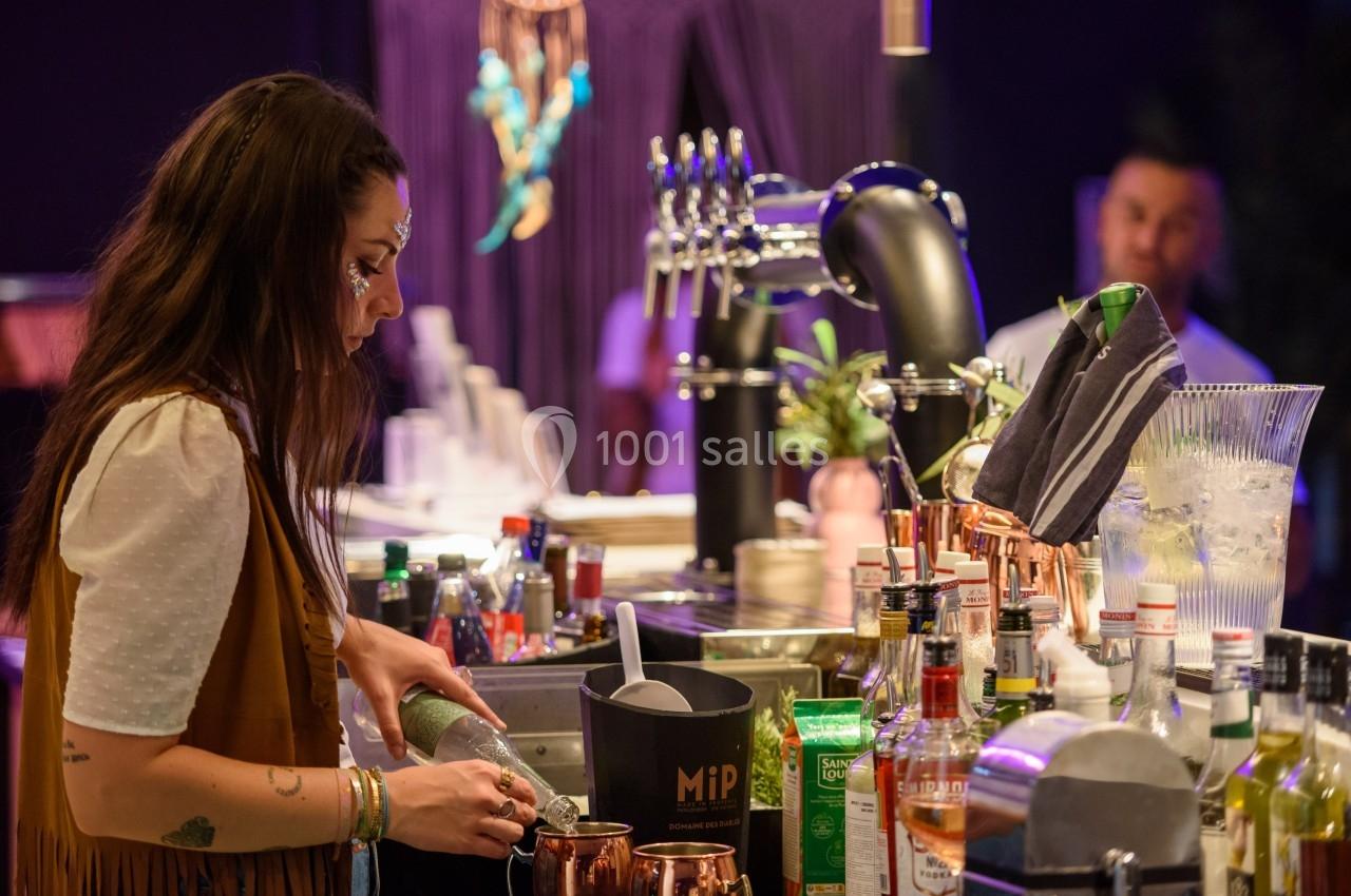 Une femme prépare des cocktails derrière un bar équipé de bouteilles, verres et tireuses à bière.