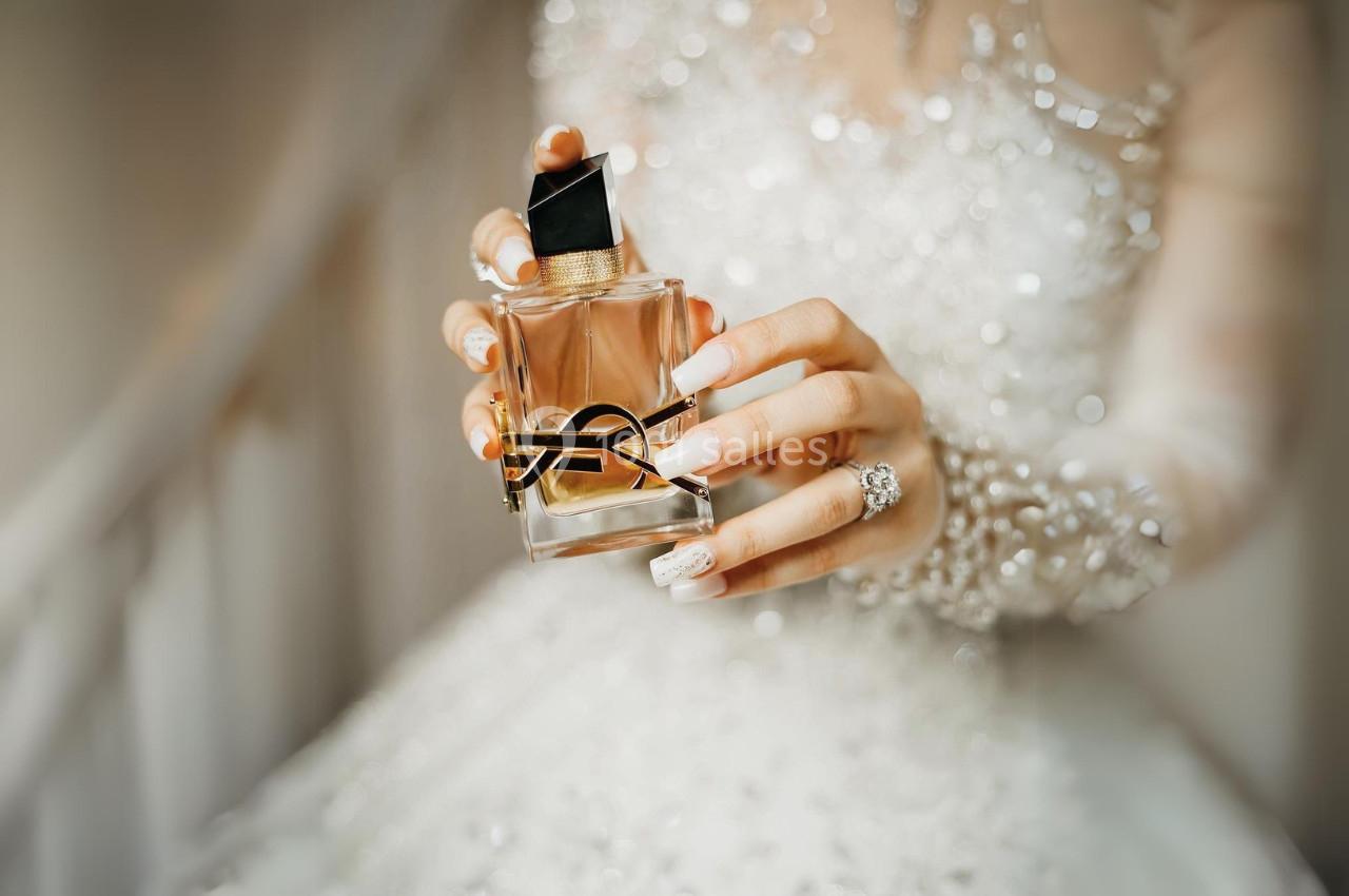 Main tenant un flacon de parfum élégant avec un design doré, en arrière-plan une robe ornée de perles.