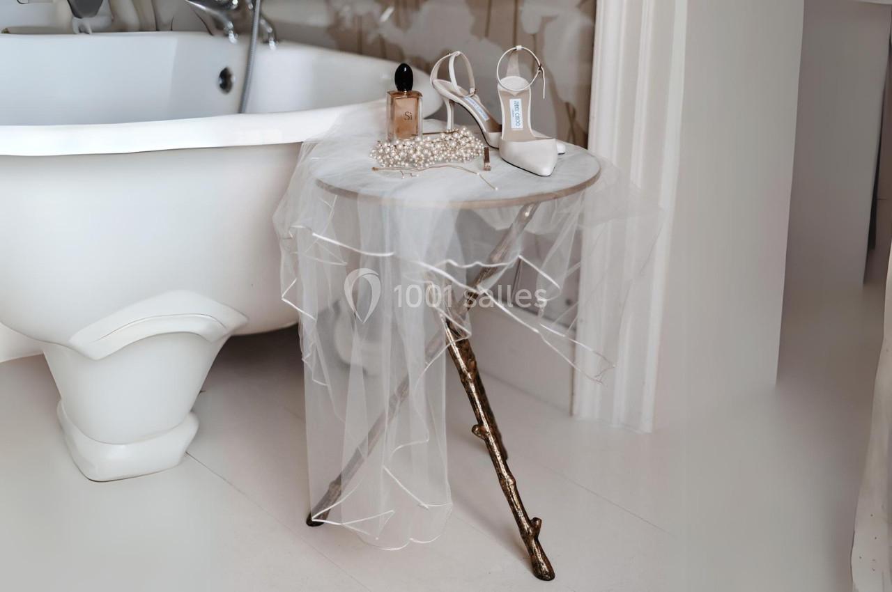 Table ronde avec voile blanc, flacon de parfum, collier de perles et chaussures à talons près d'une baignoire blanche.