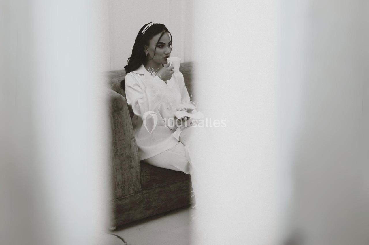 Femme assise sur un canapé, vêtue de blanc, tenant une tasse et semblant boire dans un cadre épuré.