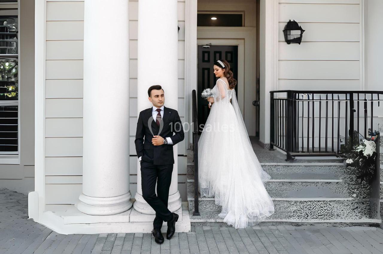Un homme en costume se tient devant un bâtiment, une femme en robe de mariée descend les marches derrière lui.
