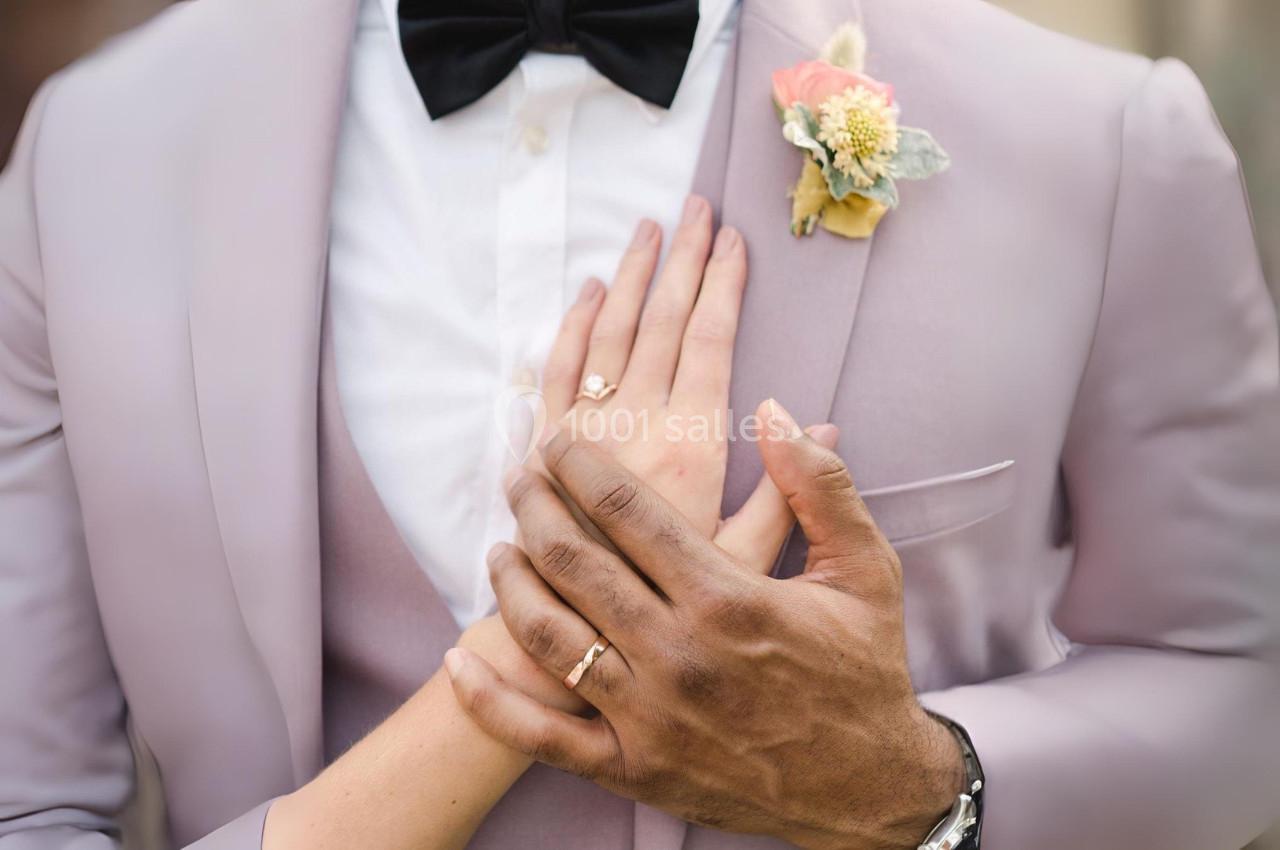 Deux mains portant des alliances entrelacées sur la poitrine d'une personne en costume rose avec une boutonnière.