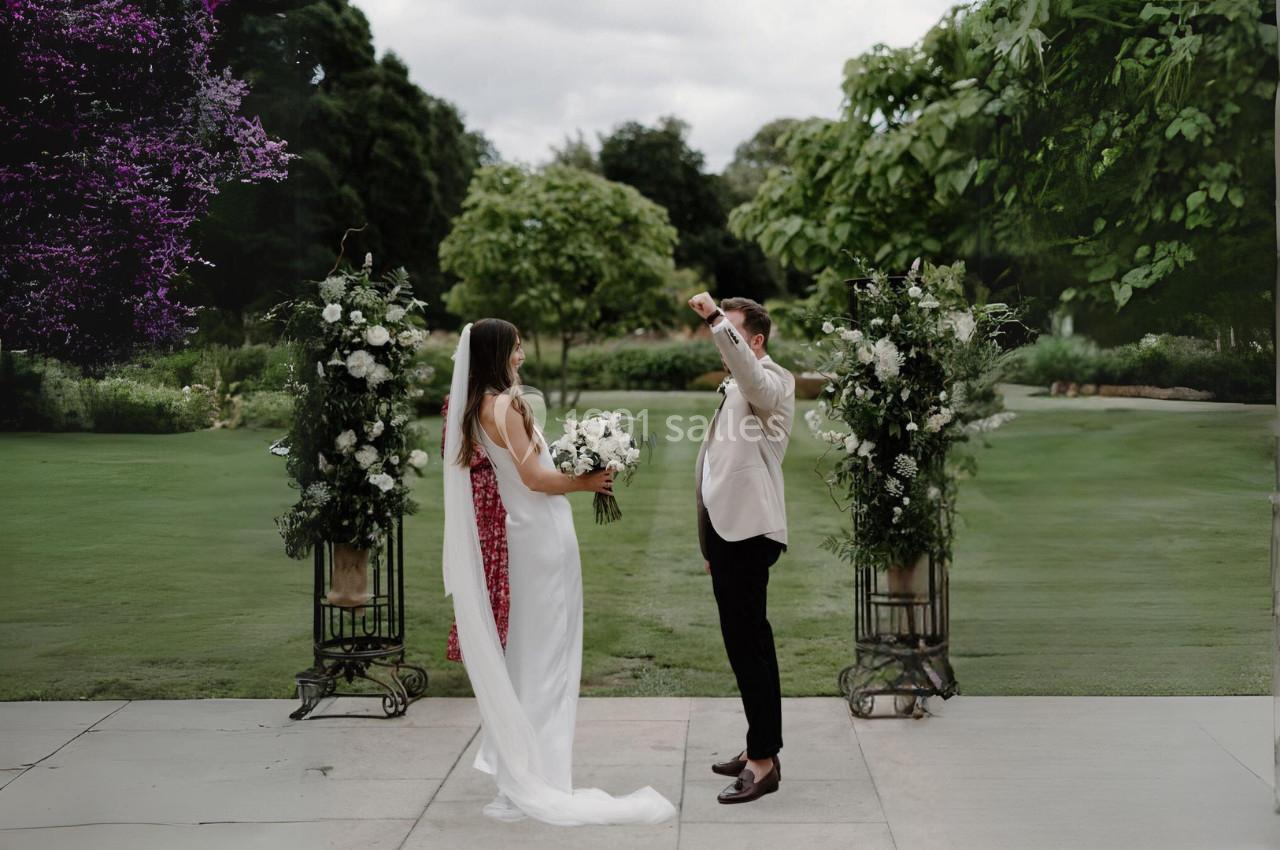 Un couple se tient face à face lors d'une cérémonie en plein air, entouré de décorations florales et d'un jardin verdoyant.