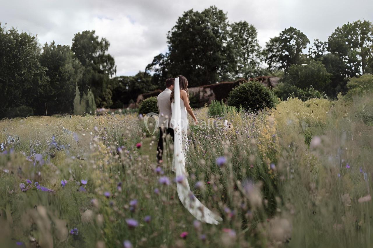 Un couple de dos marche dans un champ fleuri, entouré d'herbes hautes et d'arbres en arrière-plan.