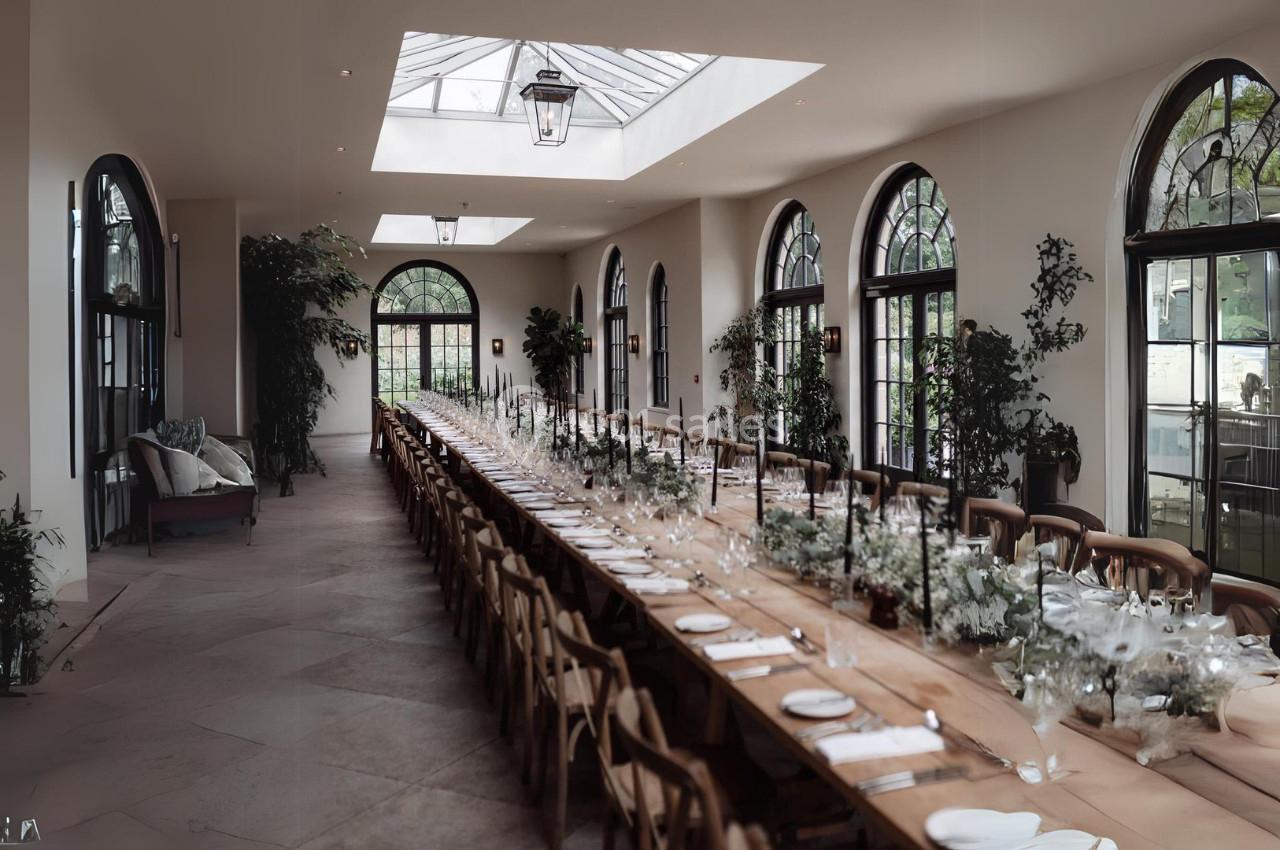 Grande table en bois dressée pour un repas dans une salle lumineuse avec verrière, arches vitrées et plantes décoratives.