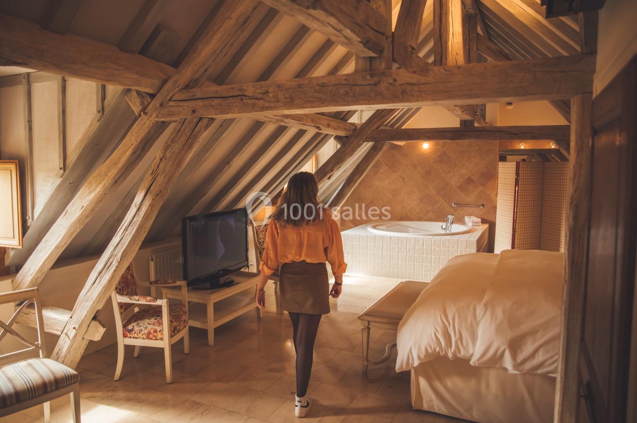 Femme de dos dans une chambre mansardée avec poutres apparentes, lit, télévision et baignoire en arrière-plan.