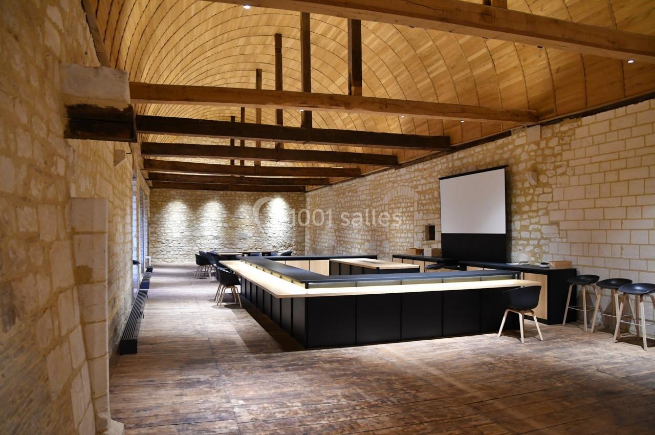 Salle avec murs en pierre, plafond voûté en bois, comptoir central moderne et chaises noires sur un sol en bois.