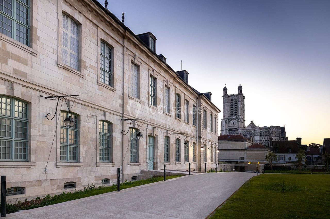 Façade d'un bâtiment historique en pierre avec des fenêtres à carreaux, vue au crépuscule, une église en arrière-plan.