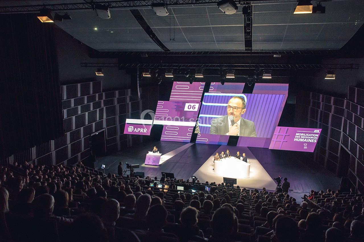 Conférence dans un auditorium avec un intervenant sur scène, écran géant et public assis dans une salle sombre.