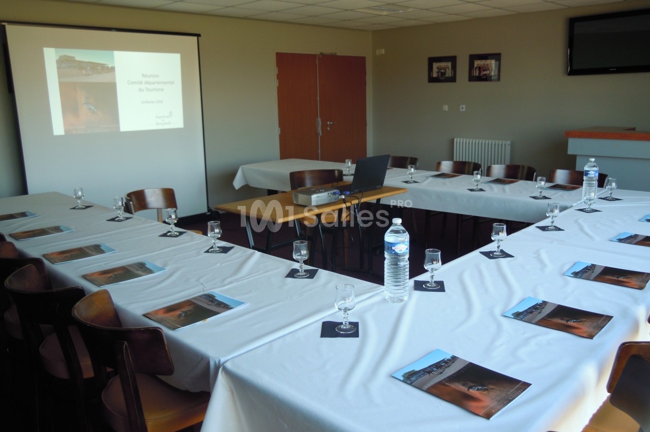 Salle de réunion aménagée en U avec des tables couvertes de nappes blanches, un projecteur et des documents disposés.