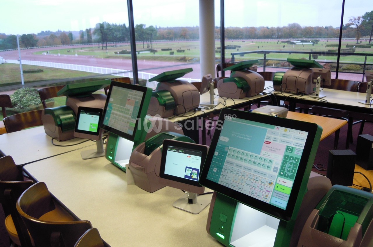 Des terminaux de paris électroniques alignés sur des tables dans une salle avec vue sur un hippodrome.