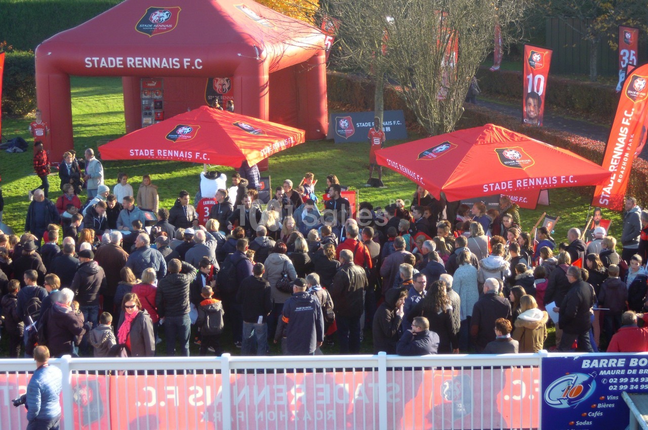 Foule rassemblée devant des stands rouges du Stade Rennais F.C. dans un espace extérieur ensoleillé.