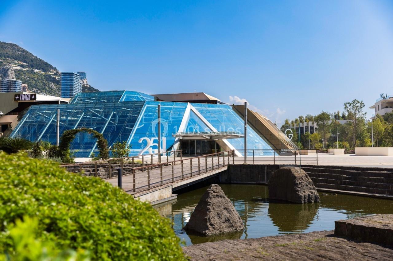 Bâtiment moderne en verre avec une passerelle en bois au-dessus d'un bassin, entouré de verdure et de rochers.