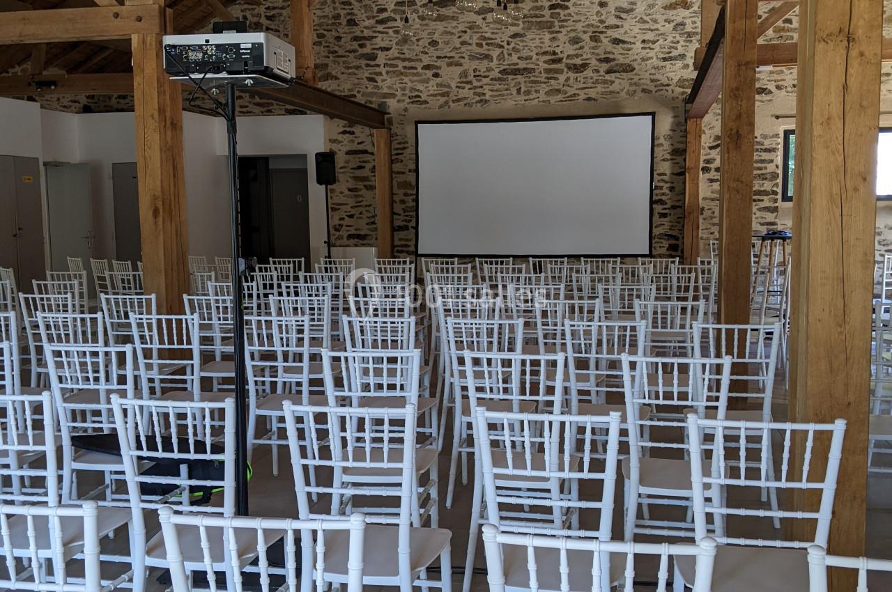 Salle de conférence avec des rangées de chaises blanches face à un écran de projection, sous une charpente en bois.