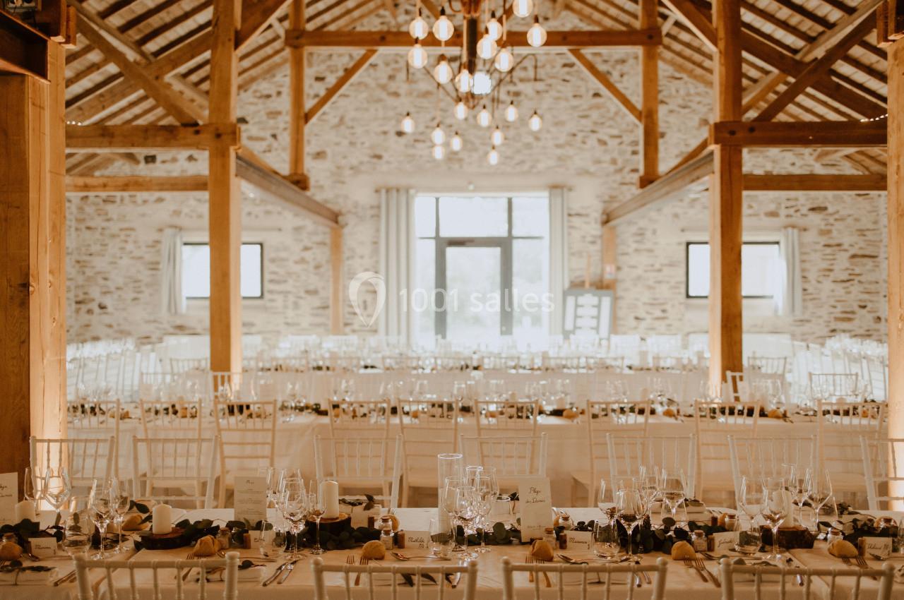 Salle de réception lumineuse avec des tables décorées, des chaises blanches et des poutres en bois apparentes.
