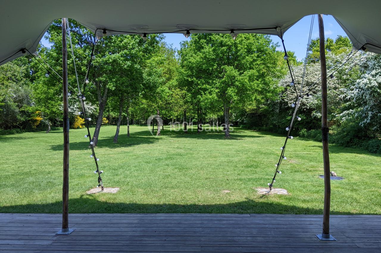 Vue d'un parc verdoyant avec pelouse, arbres et ciel dégagé, encadrée par une structure de tente ouverte.