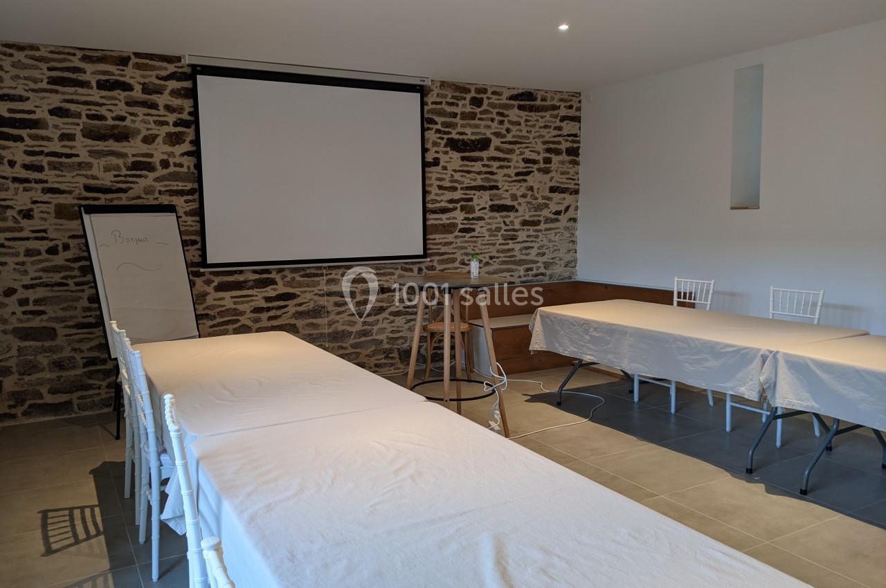 Salle de réunion avec murs en pierre, tables couvertes de nappes blanches, un tableau et un écran de projection.
