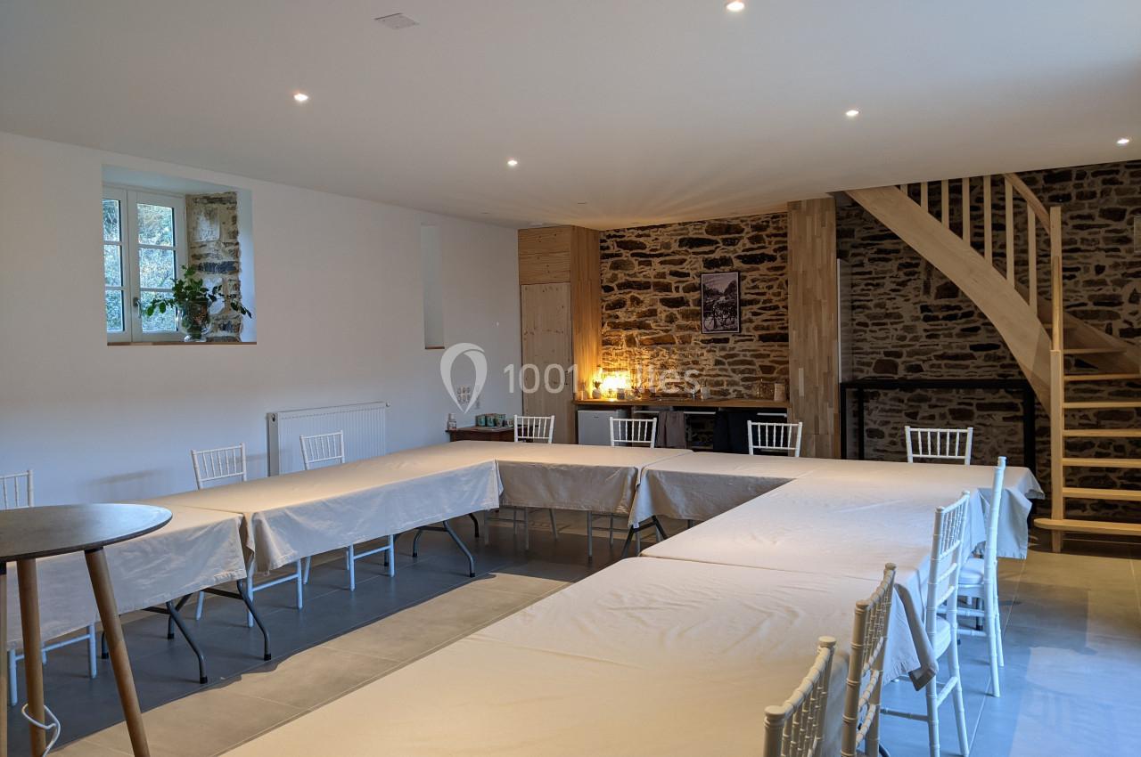Salle de réunion lumineuse avec tables disposées en U, murs en pierre apparente et escalier en bois.