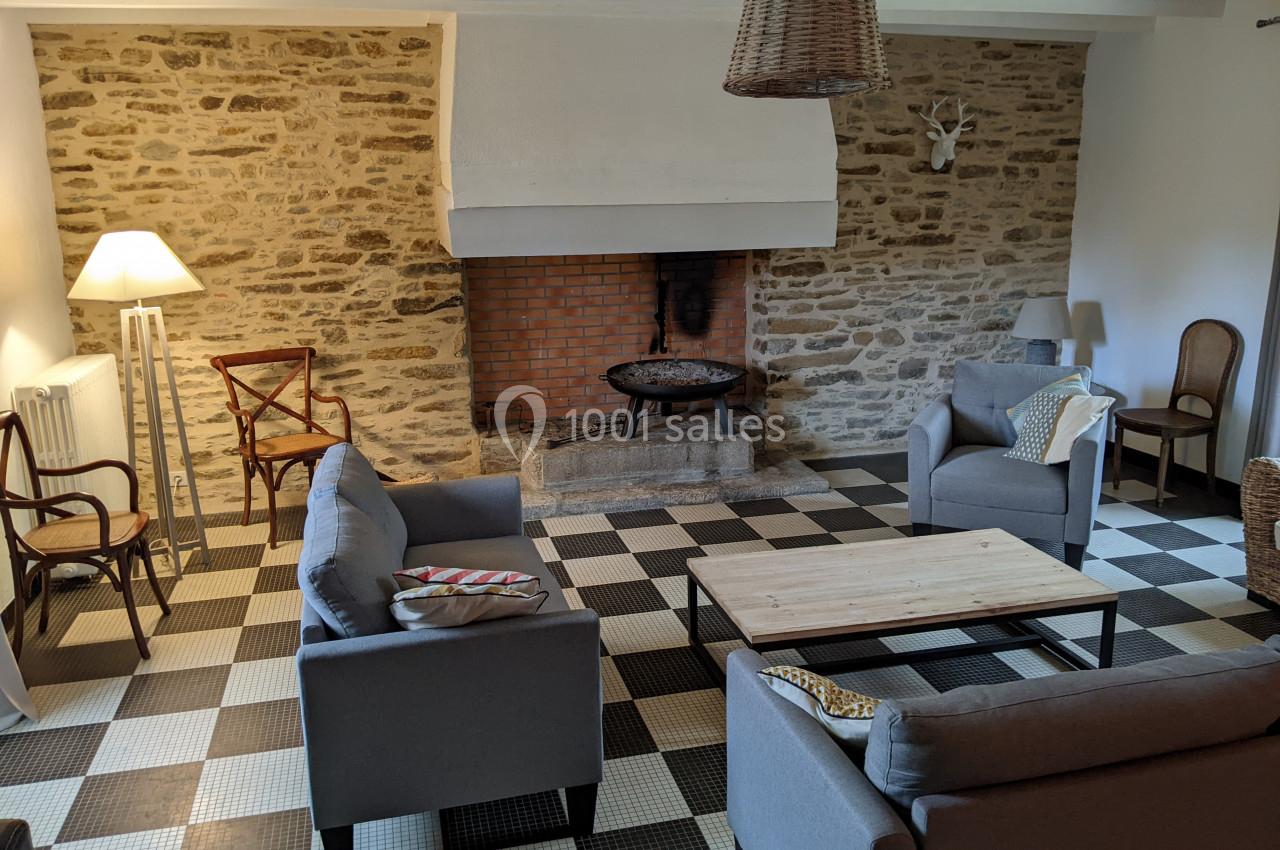 Salon avec murs en pierre, cheminée en briques, mobilier en bois et fauteuils gris sur un sol à carreaux noirs et blancs.